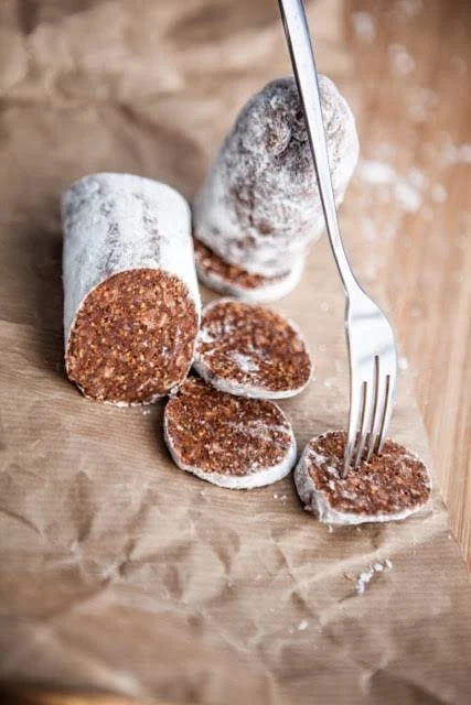 A log of fig salami dusted with flour, part-sliced into rounds on brown paper with a fork